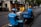 Two delivery workers wearing blue uniforms and helmets are seen on scooters, parked on the side of a city street. A person in a yellow shirt is interacting with them, possibly handing over or receiving a delivery item. The background shows a sidewalk lined with trees and storefronts.