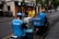 Two delivery workers wearing blue uniforms and helmets are seen on scooters, parked on the side of a city street. A person in a yellow shirt is interacting with them, possibly handing over or receiving a delivery item. The background shows a sidewalk lined with trees and storefronts.