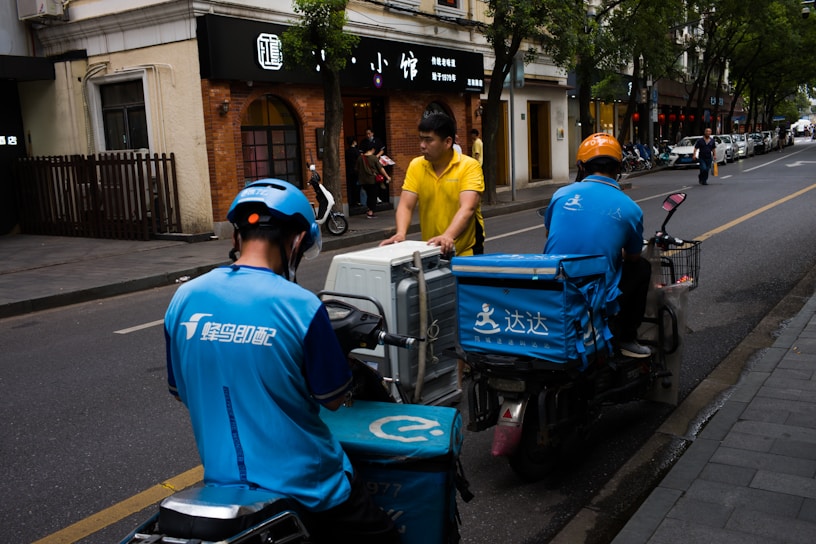 Two delivery workers wearing blue uniforms and helmets are seen on scooters, parked on the side of a city street. A person in a yellow shirt is interacting with them, possibly handing over or receiving a delivery item. The background shows a sidewalk lined with trees and storefronts.