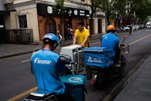 Two delivery workers wearing blue uniforms and helmets are seen on scooters, parked on the side of a city street. A person in a yellow shirt is interacting with them, possibly handing over or receiving a delivery item. The background shows a sidewalk lined with trees and storefronts.