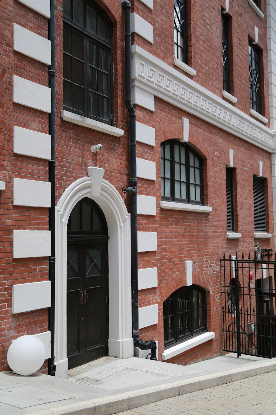 The image shows a two-story brick building with white trim and black pipes running along its exterior. The ground floor features a large arched doorway, while the upper floors have smaller windows. A white ball is placed on the sidewalk in front of the building.