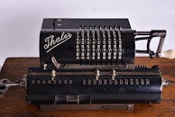A vintage mechanical calculator with a black metal body, featuring rows of numbers and levers for manual calculations. The brand 'Thales' is prominently displayed in elegant script on the top section. The device rests on a wooden surface, showcasing its intricate design and historical significance.
