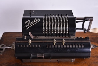 A vintage mechanical calculator with a black metal body, featuring rows of numbers and levers for manual calculations. The brand 'Thales' is prominently displayed in elegant script on the top section. The device rests on a wooden surface, showcasing its intricate design and historical significance.