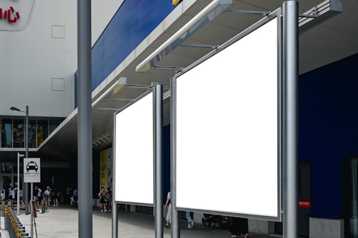 Two large blank billboards stand outside a modern building with a blue and gray facade. A few people are walking nearby, and a road sign indicating vehicle access is visible. The setting is urban, possibly near a shopping area.