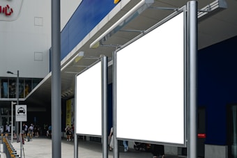 Two large blank billboards stand outside a modern building with a blue and gray facade. A few people are walking nearby, and a road sign indicating vehicle access is visible. The setting is urban, possibly near a shopping area.