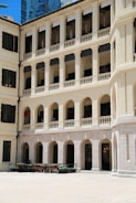 A multi-story building features colonial architecture with arched windows and balconies. The facade is cream-colored, and the windows have black shutters. The courtyard is wide with a few benches and potted plants.