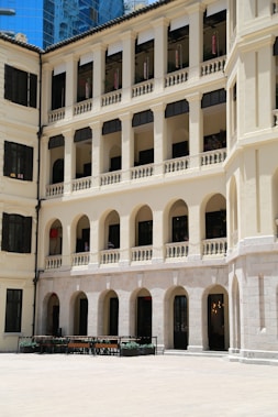 A multi-story building features colonial architecture with arched windows and balconies. The facade is cream-colored, and the windows have black shutters. The courtyard is wide with a few benches and potted plants.