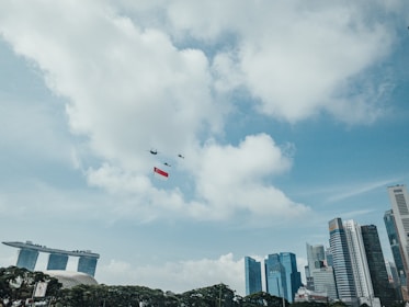 Numerous skyscrapers and modern buildings stand prominently against a partly cloudy sky. In the sky, a group of helicopters is flying in formation while carrying a large red and white flag. The iconic Marina Bay Sands hotel is visible on the left side of the image.
