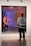 A person wearing casual clothing stands in front of a large, framed artwork. The artwork features a distorted reflection effect, creating a surreal visual. The floor is polished, and the setting appears to be a modern art gallery.