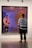 A person wearing casual clothing stands in front of a large, framed artwork. The artwork features a distorted reflection effect, creating a surreal visual. The floor is polished, and the setting appears to be a modern art gallery.