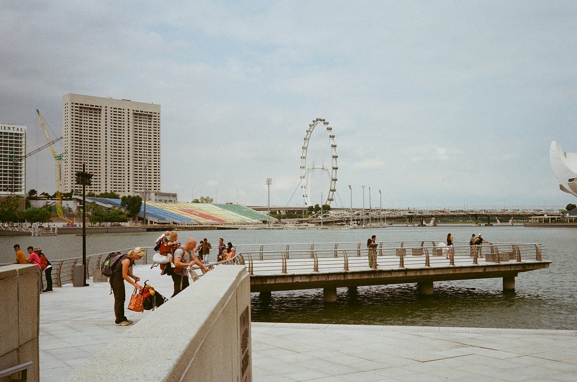 A waterfront scene with people walking and taking photographs near a river. A large Ferris wheel is visible in the background along with a colorful grandstand and modern high-rise buildings. The sky is partly cloudy, and there is a calm, relaxed atmosphere.