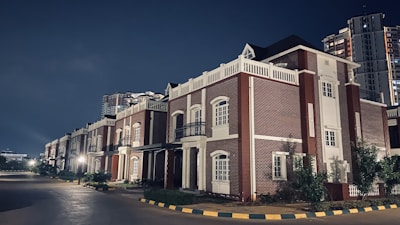 A row of elegant, modern houses featuring red brick architecture with white accents. The street is quiet and well-lit, lined with small trees and shrubs. In the background, a tall apartment building is visible against the night sky.
