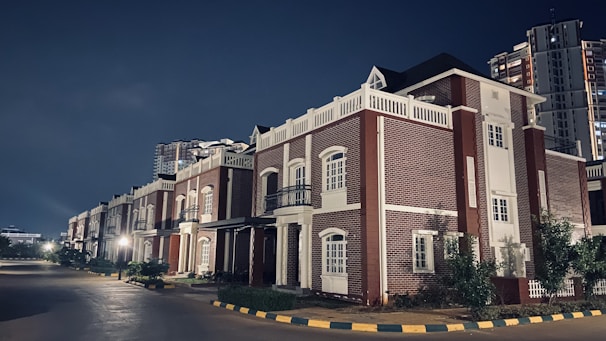 A row of elegant, modern houses featuring red brick architecture with white accents. The street is quiet and well-lit, lined with small trees and shrubs. In the background, a tall apartment building is visible against the night sky.