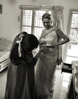 A woman is being helped with her attire, possibly a traditional Indian sari. She is smiling while another person adjusts the sari. The room has a bed, a window with curtains, and a cabinet with an open drawer.