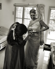 A woman is being helped with her attire, possibly a traditional Indian sari. She is smiling while another person adjusts the sari. The room has a bed, a window with curtains, and a cabinet with an open drawer.