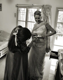 Hands skillfully draping a bright saree on a smiling woman in a sunlit room.