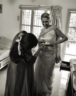 A woman is being helped with her attire, possibly a traditional Indian sari. She is smiling while another person adjusts the sari. The room has a bed, a window with curtains, and a cabinet with an open drawer.