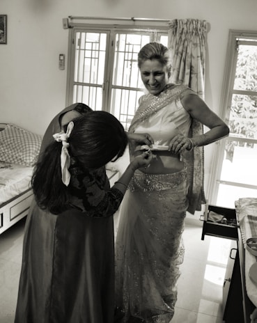 A woman is being helped with her attire, possibly a traditional Indian sari. She is smiling while another person adjusts the sari. The room has a bed, a window with curtains, and a cabinet with an open drawer.