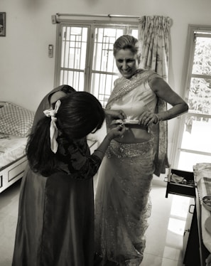 A woman is being helped with her attire, possibly a traditional Indian sari. She is smiling while another person adjusts the sari. The room has a bed, a window with curtains, and a cabinet with an open drawer.
