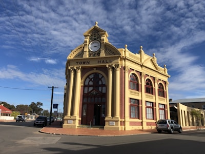 A historic town hall building with an ornate facade featuring a large central clock and arched windows. The building is constructed with red bricks and cream stone accents, embodying classic architectural elements. The clear sky is dotted with white clouds, and there are a few cars parked on the surrounding streets.