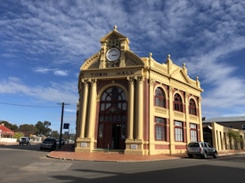 A historic town hall building with an ornate facade featuring a large central clock and arched windows. The building is constructed with red bricks and cream stone accents, embodying classic architectural elements. The clear sky is dotted with white clouds, and there are a few cars parked on the surrounding streets.