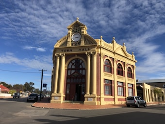 A historic town hall building with an ornate facade featuring a large central clock and arched windows. The building is constructed with red bricks and cream stone accents, embodying classic architectural elements. The clear sky is dotted with white clouds, and there are a few cars parked on the surrounding streets.