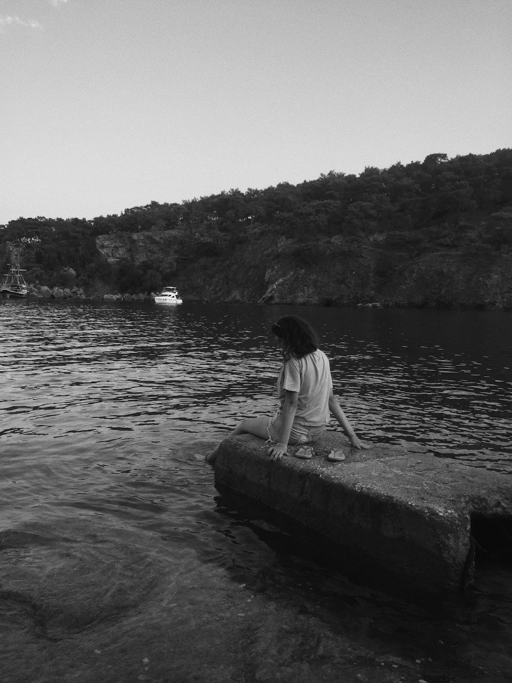 grayscale photo of woman in white shirt sitting on rock in the middle of the lake