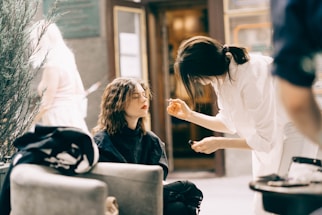 A makeup artist is applying cosmetics to a woman seated in an outdoor setting. The woman has shoulder-length wavy hair and is sitting calmly with her eyes closed, while the artist is intently focused on her work. There is greenery in the background and an open doorway, suggesting this is taking place possibly outside a salon or shop.