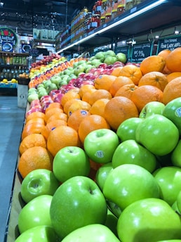 A vibrant display of fresh fruits arranged in neat rows. Green apples, oranges, and red apples are prominently featured, creating a colorful and inviting scene. Jars and bottles are stacked on shelves above, adding variety and depth. Handwritten chalkboard signs with pricing information are visible.