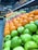 A vibrant display of fresh fruits arranged in neat rows. Green apples, oranges, and red apples are prominently featured, creating a colorful and inviting scene. Jars and bottles are stacked on shelves above, adding variety and depth. Handwritten chalkboard signs with pricing information are visible.