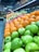 A vibrant display of fresh fruits arranged in neat rows. Green apples, oranges, and red apples are prominently featured, creating a colorful and inviting scene. Jars and bottles are stacked on shelves above, adding variety and depth. Handwritten chalkboard signs with pricing information are visible.