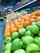 A vibrant display of fresh fruits arranged in neat rows. Green apples, oranges, and red apples are prominently featured, creating a colorful and inviting scene. Jars and bottles are stacked on shelves above, adding variety and depth. Handwritten chalkboard signs with pricing information are visible.
