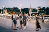 A group of people is gathered on a city street in the evening, with streetlights illuminating the scene. One person appears to be filming or taking photos, as they are holding a camera mounted on a stabilizer. A woman in a long, patterned dress and high heels is standing in profile to the group, possibly posing. Cars and trees with blurred city lights are visible in the background, creating a lively urban atmosphere.