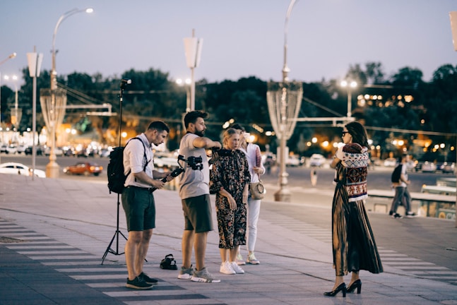 A group of people is gathered on a city street in the evening, with streetlights illuminating the scene. One person appears to be filming or taking photos, as they are holding a camera mounted on a stabilizer. A woman in a long, patterned dress and high heels is standing in profile to the group, possibly posing. Cars and trees with blurred city lights are visible in the background, creating a lively urban atmosphere.