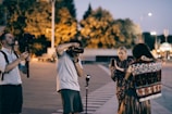 A group of people engaged in a photography session outdoors during twilight. One person is holding a large camera with a flash, another is observing with a steady camera on a stabilizer, and a third person is using a smartphone. The scene is set in an urban environment with blurred trees and streetlights in the background, suggesting evening or early night time.