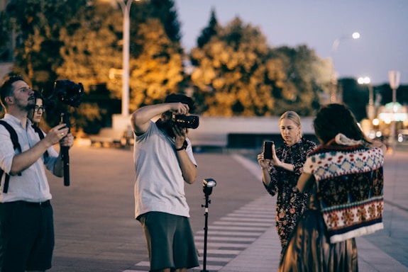 A group of people engaged in a photography session outdoors during twilight. One person is holding a large camera with a flash, another is observing with a steady camera on a stabilizer, and a third person is using a smartphone. The scene is set in an urban environment with blurred trees and streetlights in the background, suggesting evening or early night time.