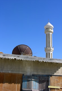 A mosque minaret rises prominently against a clear blue sky, with a rough stone wall and a rusty satellite dish visible in the foreground. The building appears weathered, with boarded windows and a corrugated roof.