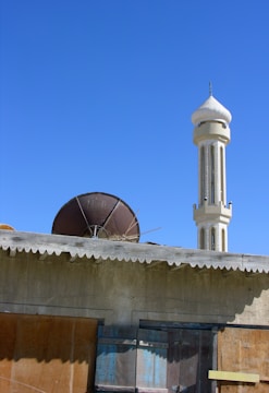 A mosque minaret rises prominently against a clear blue sky, with a rough stone wall and a rusty satellite dish visible in the foreground. The building appears weathered, with boarded windows and a corrugated roof.