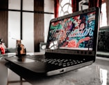 A laptop on a table with a vibrant, colorful screen displaying an artistic design. The background includes wooden cabinets and patterned fabric curtains. The table reflects the laptop, and a decorative cup sits nearby, adding a contemporary feel to the setting.