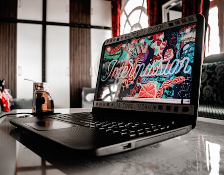 A laptop on a table with a vibrant, colorful screen displaying an artistic design. The background includes wooden cabinets and patterned fabric curtains. The table reflects the laptop, and a decorative cup sits nearby, adding a contemporary feel to the setting.