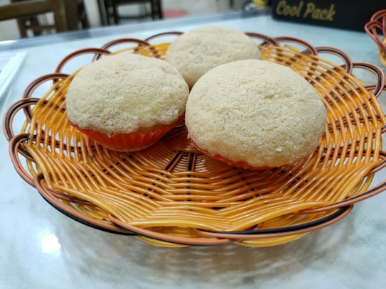 Three round, fluffy baked goods rest on an ornate, woven orange basket, placed on a marble countertop.