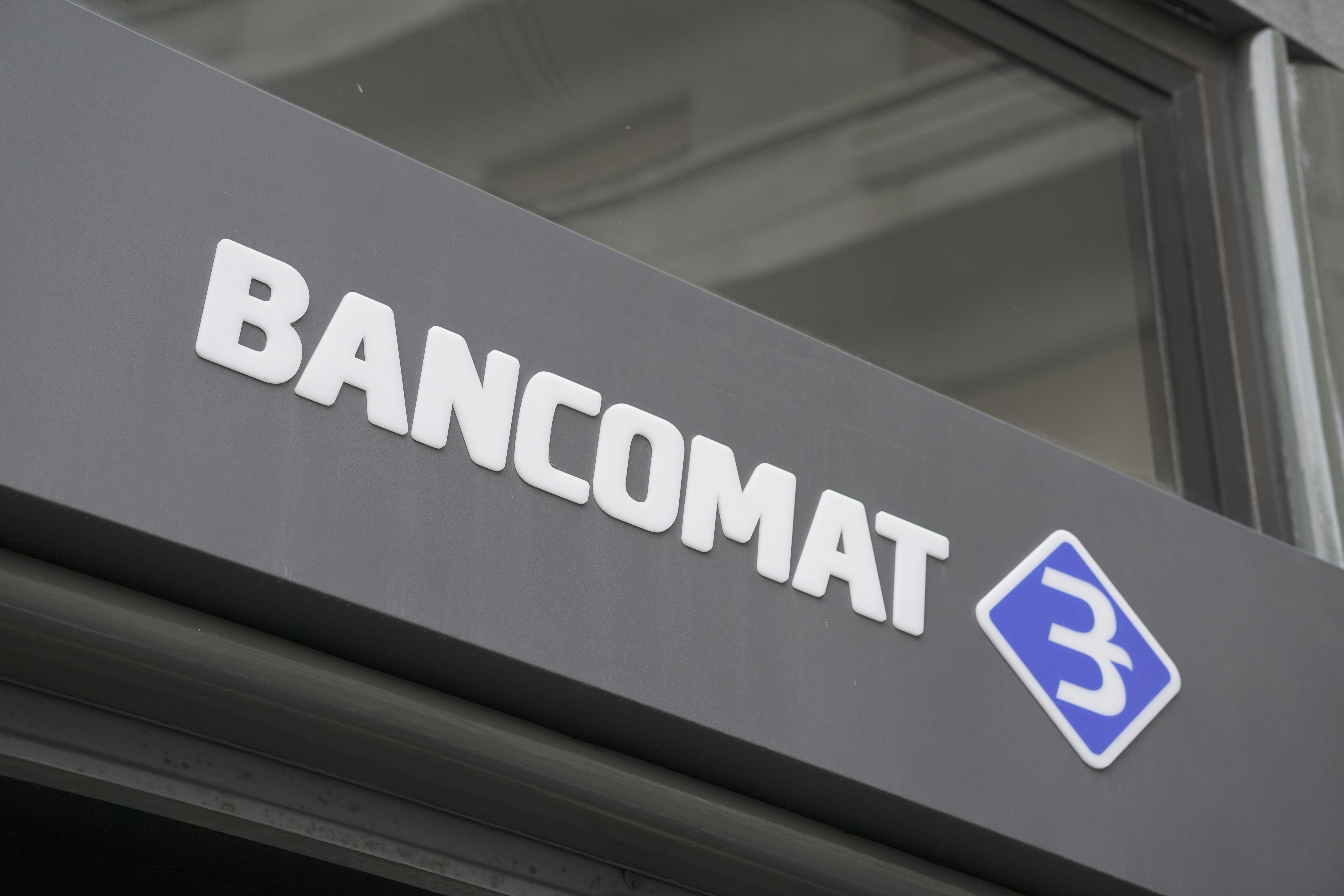 A modern sign with the word 'BANCOMAT' in bold, white letters is prominently displayed on a dark surface. To the right of the text is a blue and white logo resembling a stylized letter 'B' encased in a diamond shape.