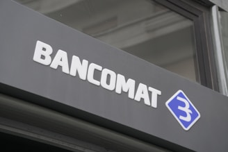 A modern sign with the word 'BANCOMAT' in bold, white letters is prominently displayed on a dark surface. To the right of the text is a blue and white logo resembling a stylized letter 'B' encased in a diamond shape.
