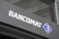 A modern sign with the word 'BANCOMAT' in bold, white letters is prominently displayed on a dark surface. To the right of the text is a blue and white logo resembling a stylized letter 'B' encased in a diamond shape.