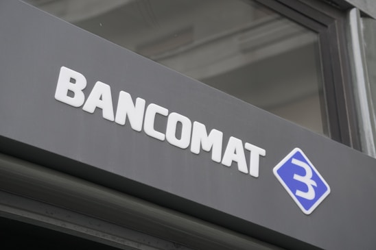 A modern sign with the word 'BANCOMAT' in bold, white letters is prominently displayed on a dark surface. To the right of the text is a blue and white logo resembling a stylized letter 'B' encased in a diamond shape.