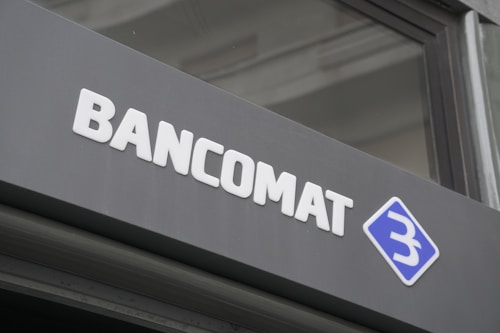 A modern sign with the word 'BANCOMAT' in bold, white letters is prominently displayed on a dark surface. To the right of the text is a blue and white logo resembling a stylized letter 'B' encased in a diamond shape.