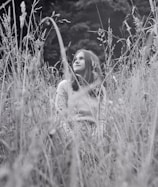A black-and-white photograph of a young person sitting amidst tall wild grasses, gazing upward. The scene conveys a sense of tranquility with soft light filtering through, accentuating the natural surroundings.