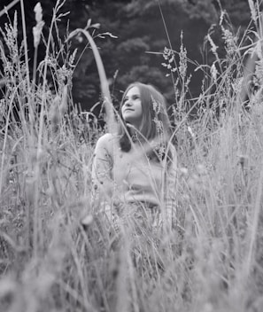 A black-and-white photograph of a young person sitting amidst tall wild grasses, gazing upward. The scene conveys a sense of tranquility with soft light filtering through, accentuating the natural surroundings.