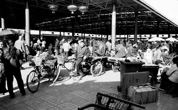 A bustling market scene filled with people engaged in various activities. Several bicycles and rickshaws are parked, and groups of people are gathered under a large canopy structure. The area is lively with individuals interacting, shopping, and moving about.