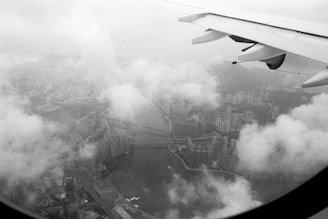 A black and white aerial view of a cityscape covered partly by clouds with an aircraft wing visible in the upper right corner of the image. Skyscrapers and a river or waterway can be seen through the gaps in the clouds.